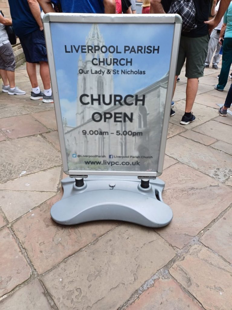 Picture shows a free-standing notice-board on the pavement. The board says "Liverpool Parish Church. Our Lady and St Nicholas. Church Open 9am to 5pm