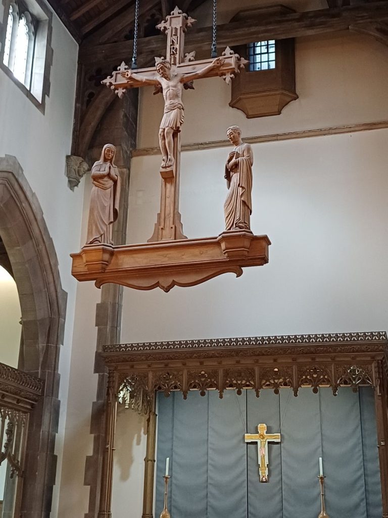 Picture shows a large wooden carving of Jesus on the cross, with Mary and John either side. The carving is suspended from the roof