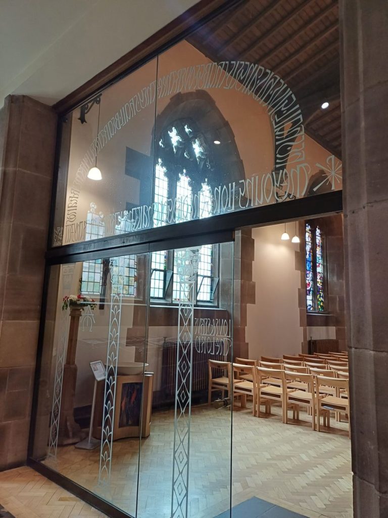 Picture shows glass dors and screen, both etched with writing and patterns. Beyond the screen are rows of chairs at the back of a church building. 