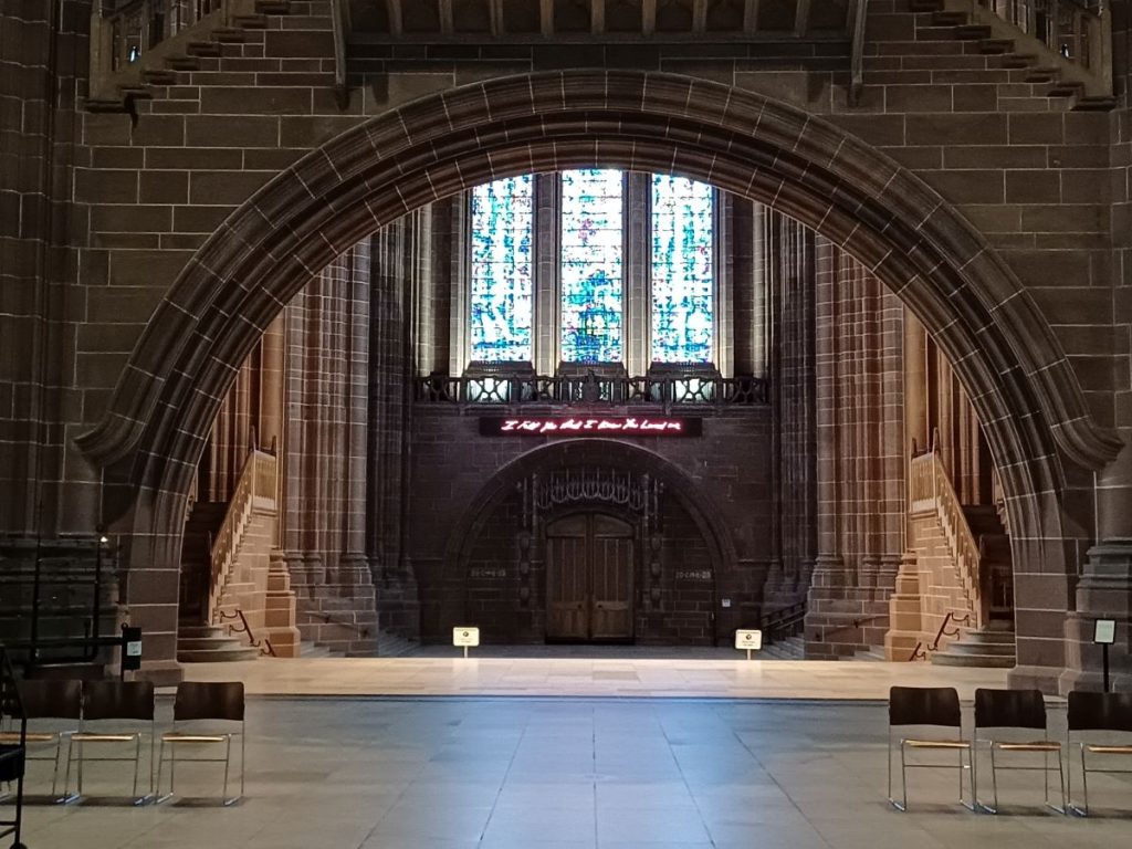 Picture shows the interior of Liverpool Cathedral looking through a large stone archway towards the West doors. Above the doors is written in neon lights "I felt you and I knew the Lord"
