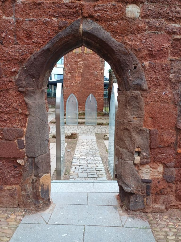 Picture shows a pointed arch in a red-stone wall, through which you can see a gravel path and some glass tombstone shapes.
