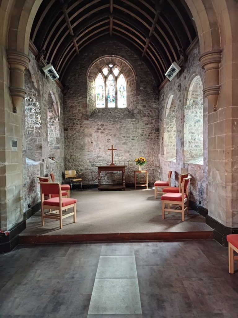 Image shows the interior of a small, ancient church. The space is open, with a few chairs arranged in a loose circle. In the background is an altar with a cross on it. 
