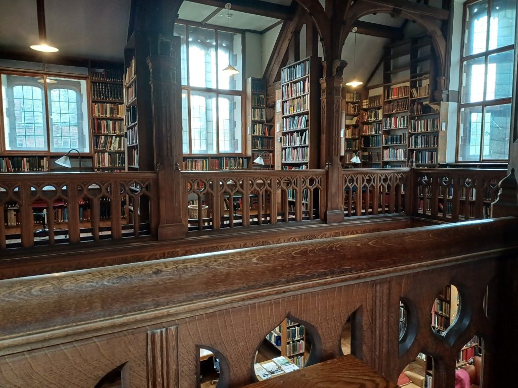 Picture shows a Victorian library. It is taken from a first floor gallery, looking across to the gallery on the other side. Tall shelves of books separate reading desks.