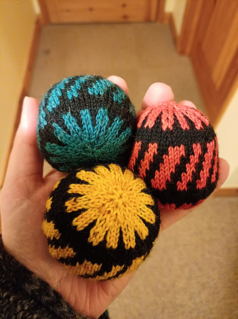 Picture shows three knitted balls, held in a hand against a blurred background. The knitting is two-colour stranded, each ball using black as the main colour and then either red, blue or yellow for the contrast.